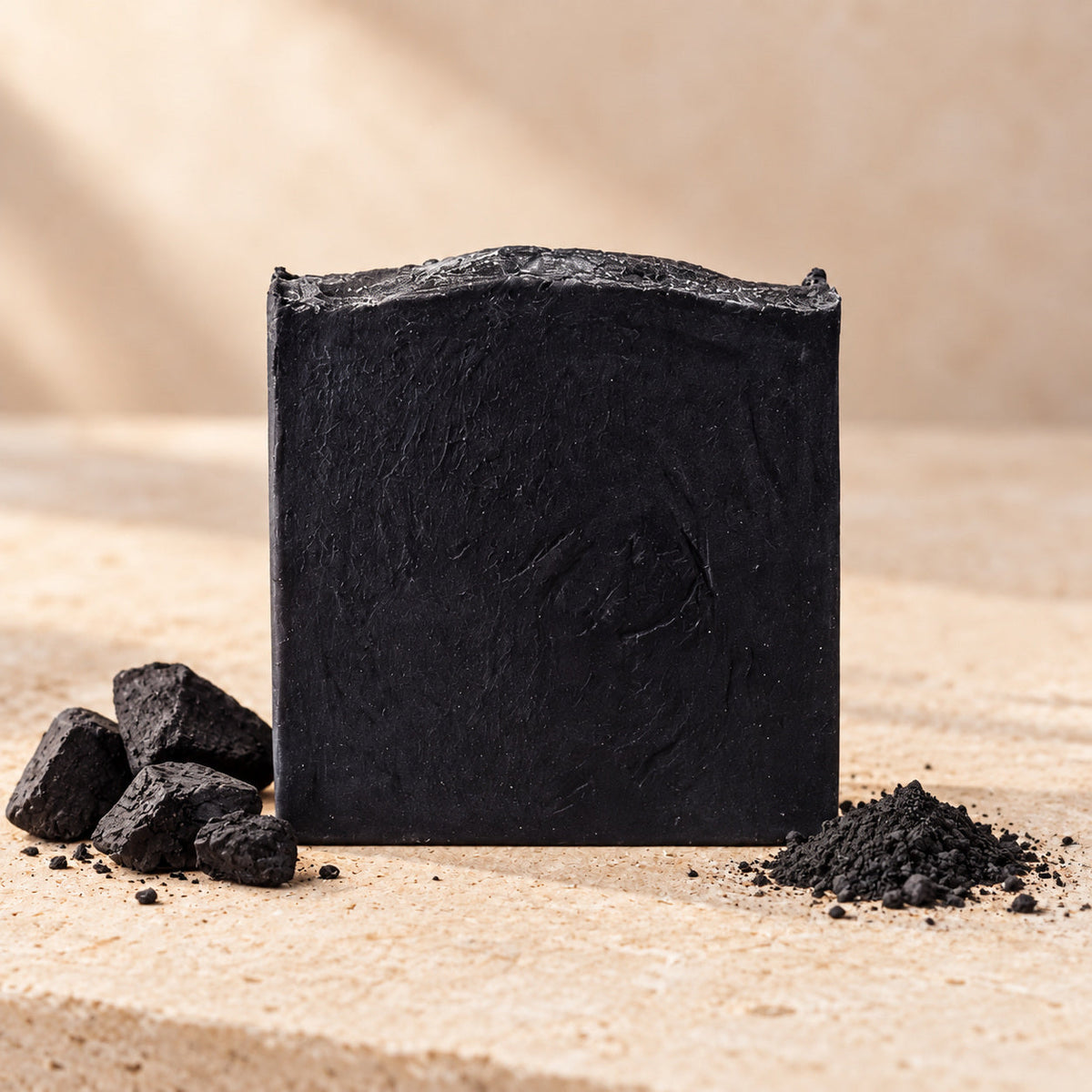 Jabón de carbón activado de maro naturals, barra negra junto a trozos y polvo de carbón sobre piedra clara.