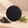 Shampoo sólido de carbón y árbol de té de maro naturals, barra negra junto a carbón y hojas finas de árbol de té.